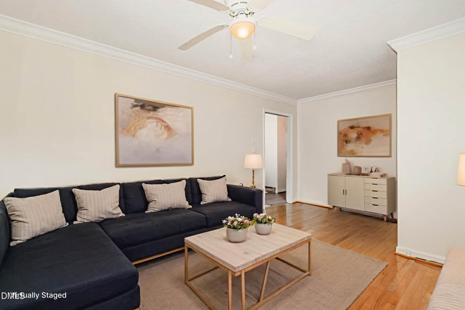 A bright living room with a dark sectional sofa, beige cushions, a wooden coffee table, potted plants, wall art, a sideboard, hardwood floors, and a ceiling fan. Virtually staged furniture and decor are visible.