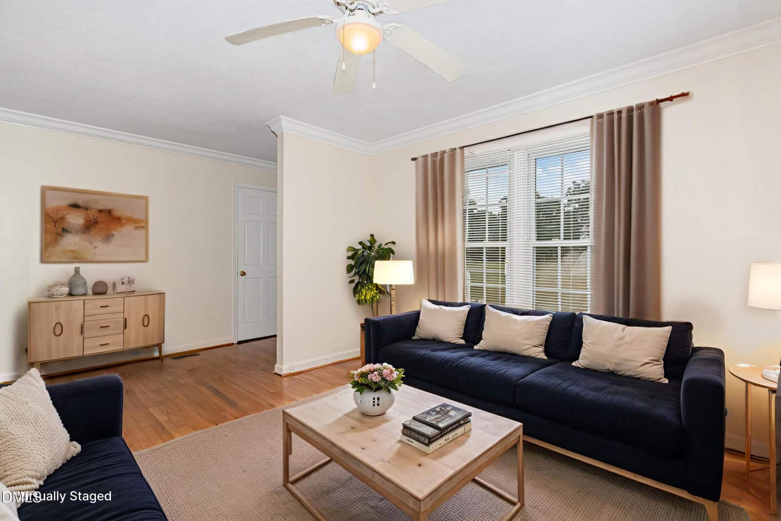 A bright living room with a navy sofa, beige pillows, a coffee table with books and flowers, a sideboard, wall art, potted plant, and large window with brown curtains. Ceiling fan and wood floor are visible.