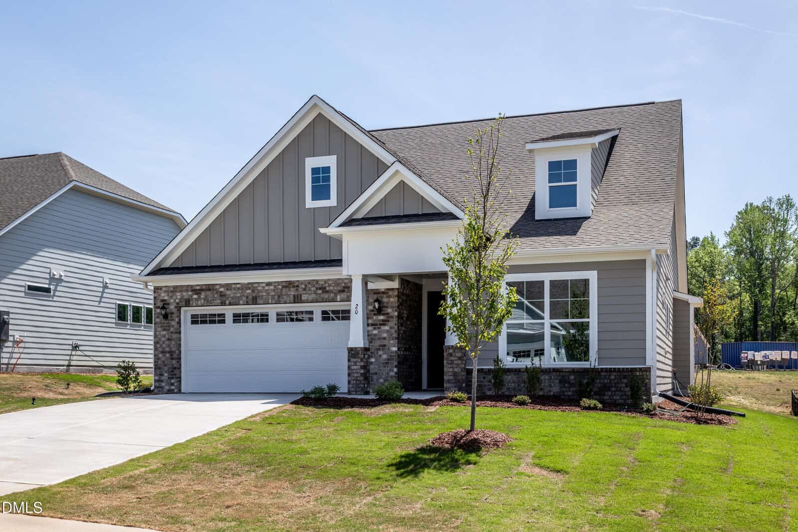 A modern two-story suburban house with gray siding, brick accents, a front porch, and attached two-car garage. The yard is freshly landscaped with green grass and a small tree in front.