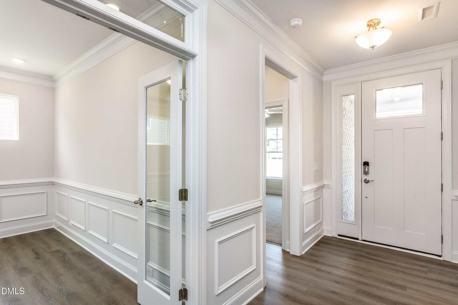 Bright entryway of a house with white walls, decorative wainscoting, glass-paned office doors, wood flooring, and a front door with sidelights. Natural light fills the space through large windows.