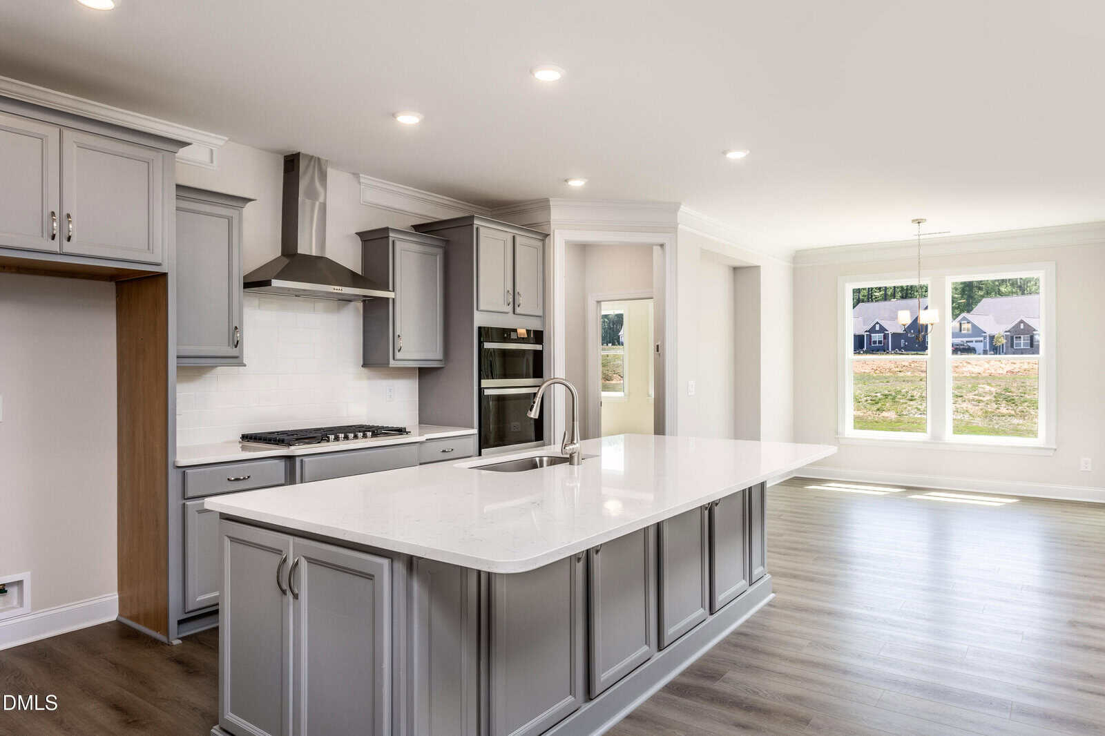 A modern kitchen with gray cabinets, a white island with a built-in sink, stainless steel appliances, and large windows providing natural light into the adjacent dining area.