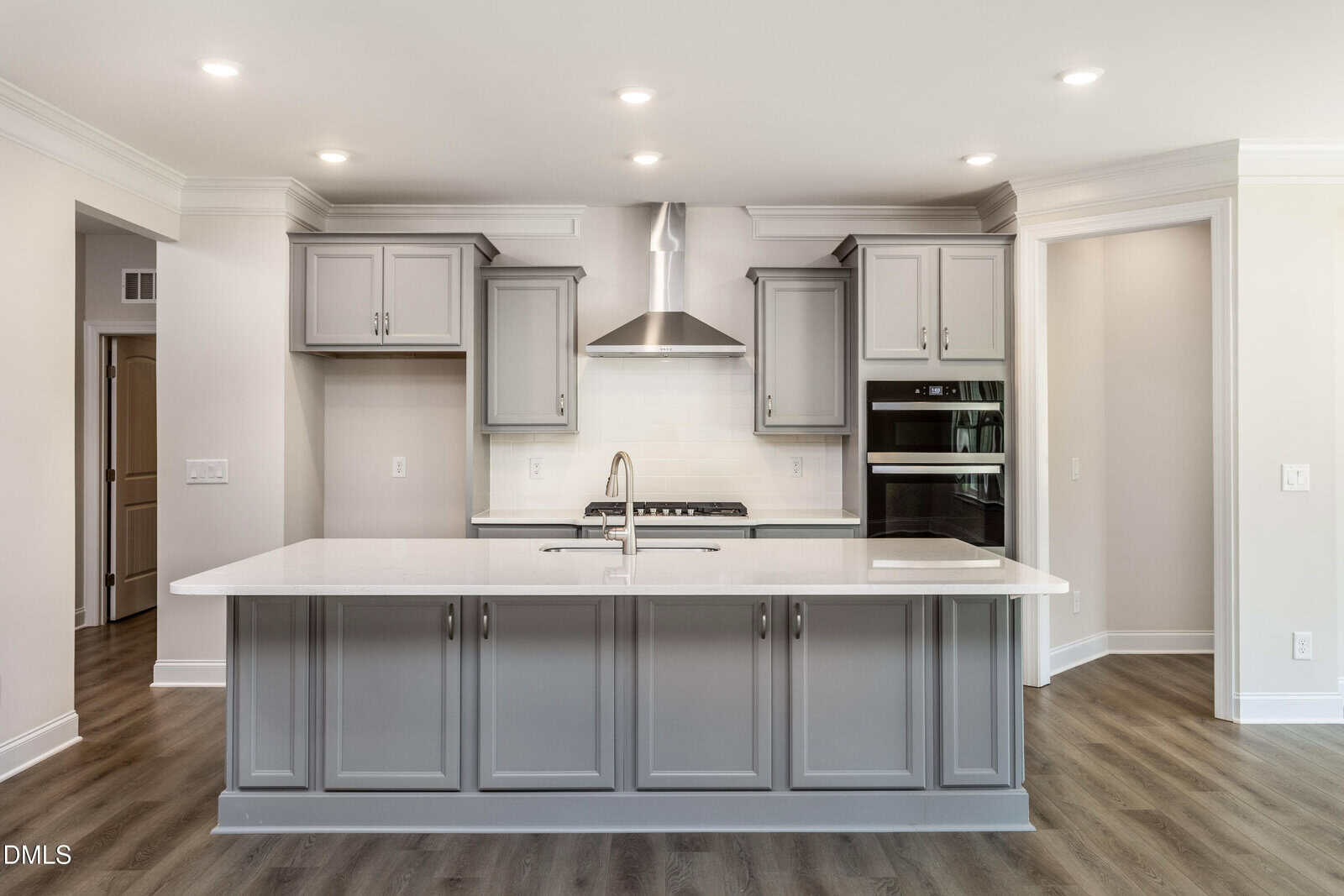 A modern kitchen features gray cabinets, stainless steel appliances, a range hood, double wall ovens, a white tile backsplash, an island with a sink, and wood-style flooring under recessed ceiling lights.