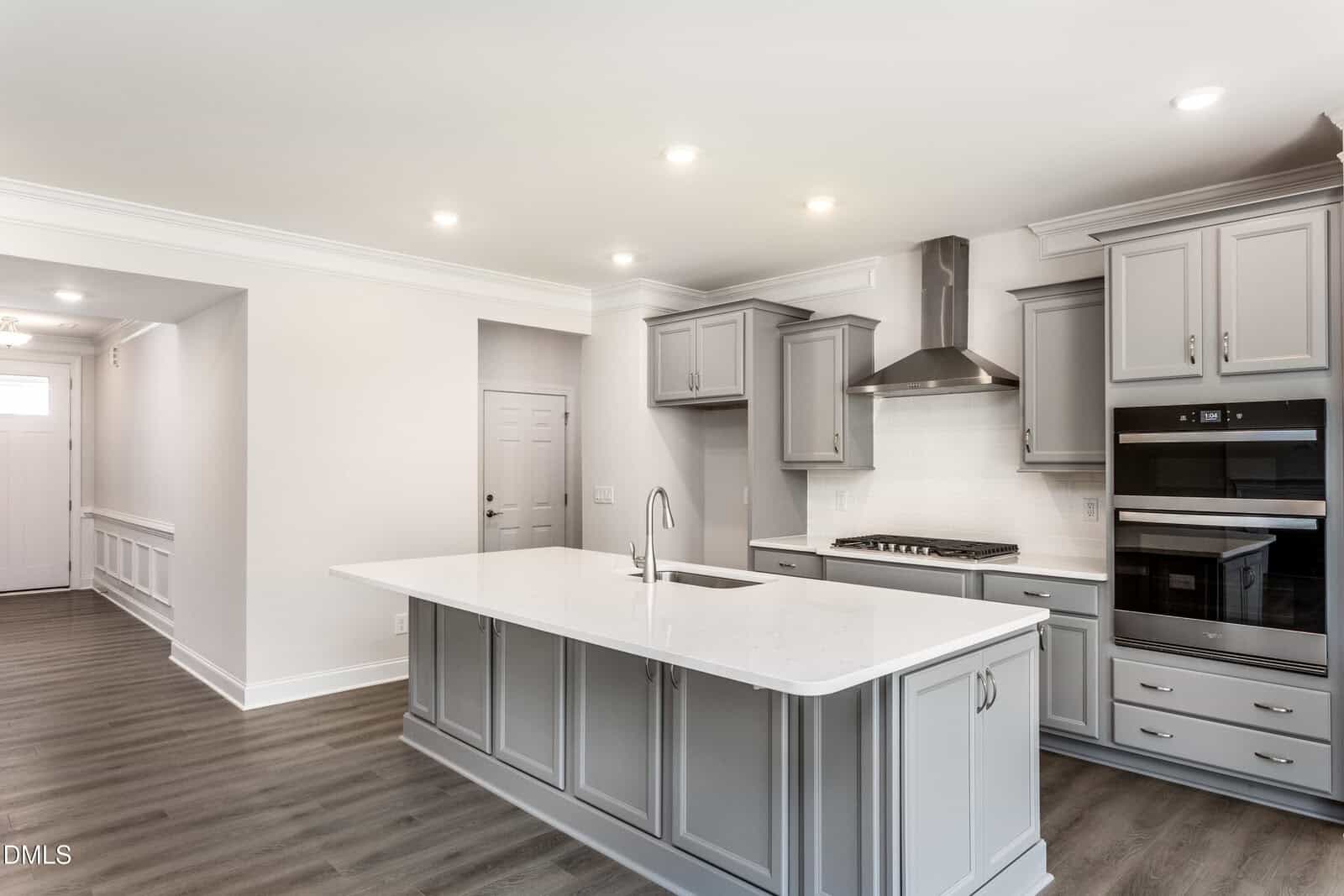 Modern kitchen with gray cabinets, a white island countertop, stainless steel appliances, built-in oven, stove with hood, wood flooring, and recessed lighting. White walls and ceilings create a bright, clean space.