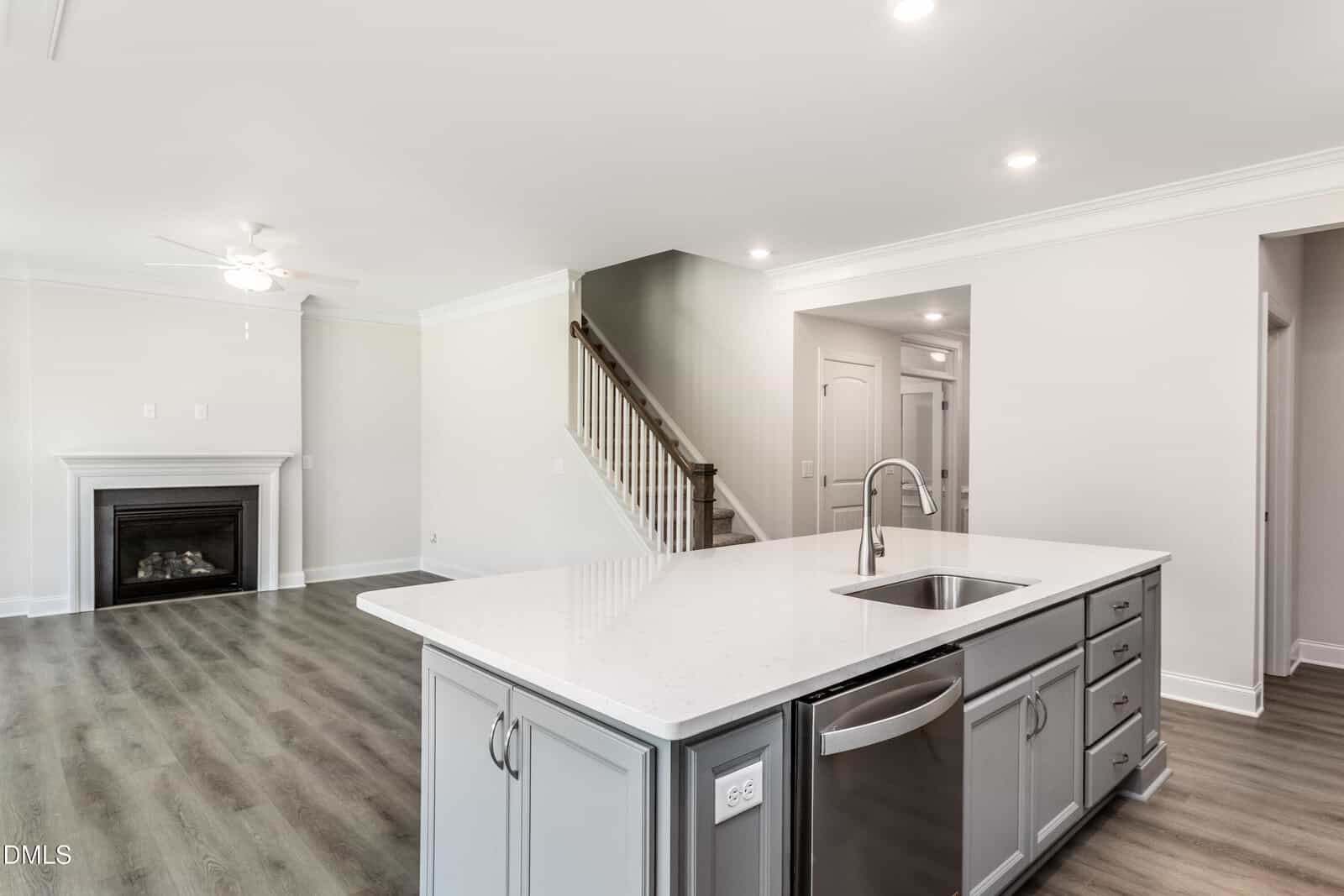 A modern open-concept kitchen with a white island, stainless steel sink, gray cabinets, and a view of a living area with a fireplace, ceiling fan, and staircase. Walls are light and floors are wood-look.