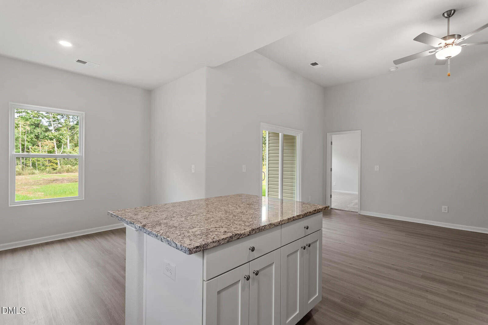 A modern, unfurnished open-concept living area with light gray walls, wood-look flooring, a ceiling fan, and a kitchen island with a granite countertop and white cabinets. Sunlight enters through two windows.