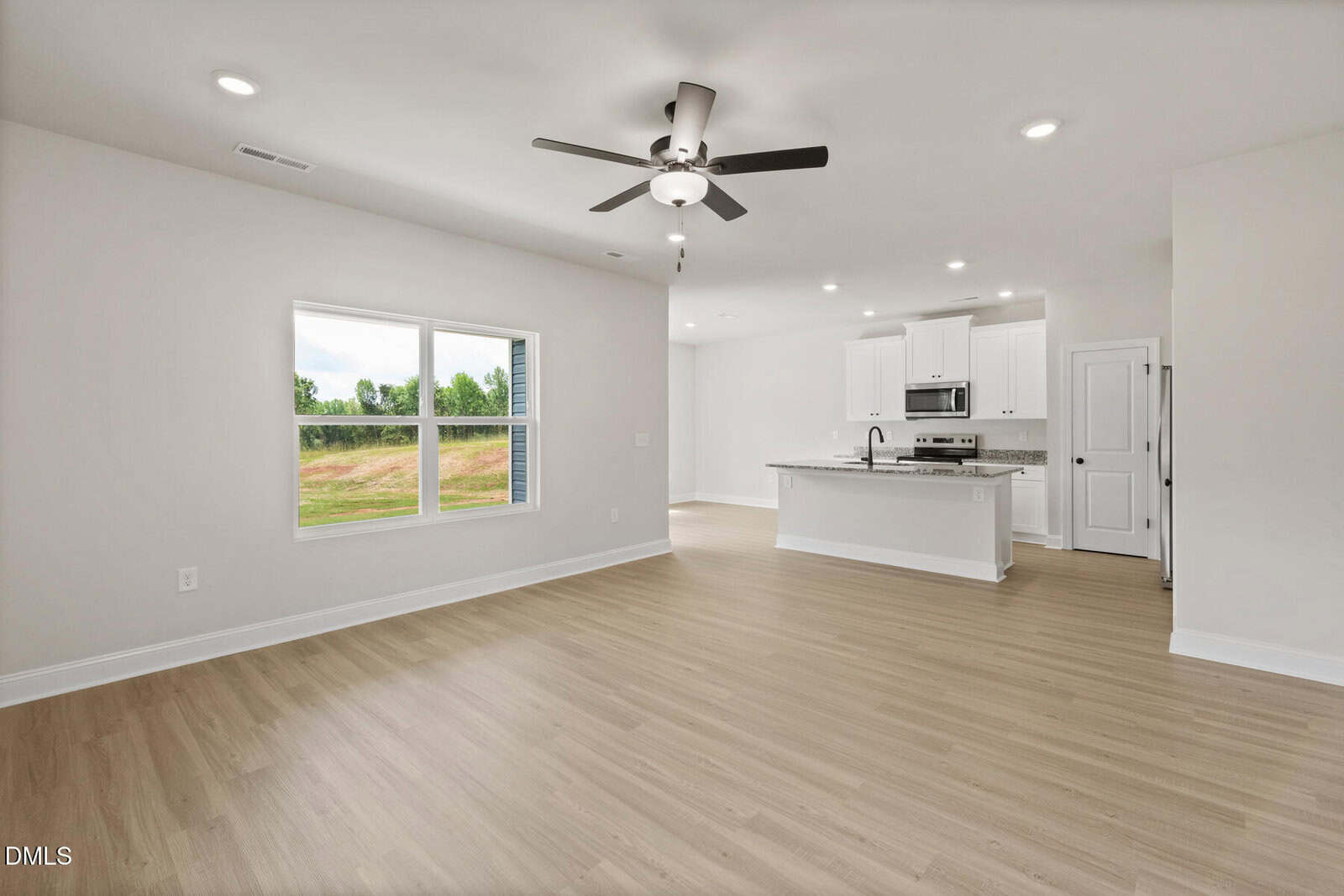 Unfurnished open-plan living area with light wood flooring, white walls, a ceiling fan with lights, a large window, and a kitchen featuring white cabinets, stainless steel appliances, and a center island.