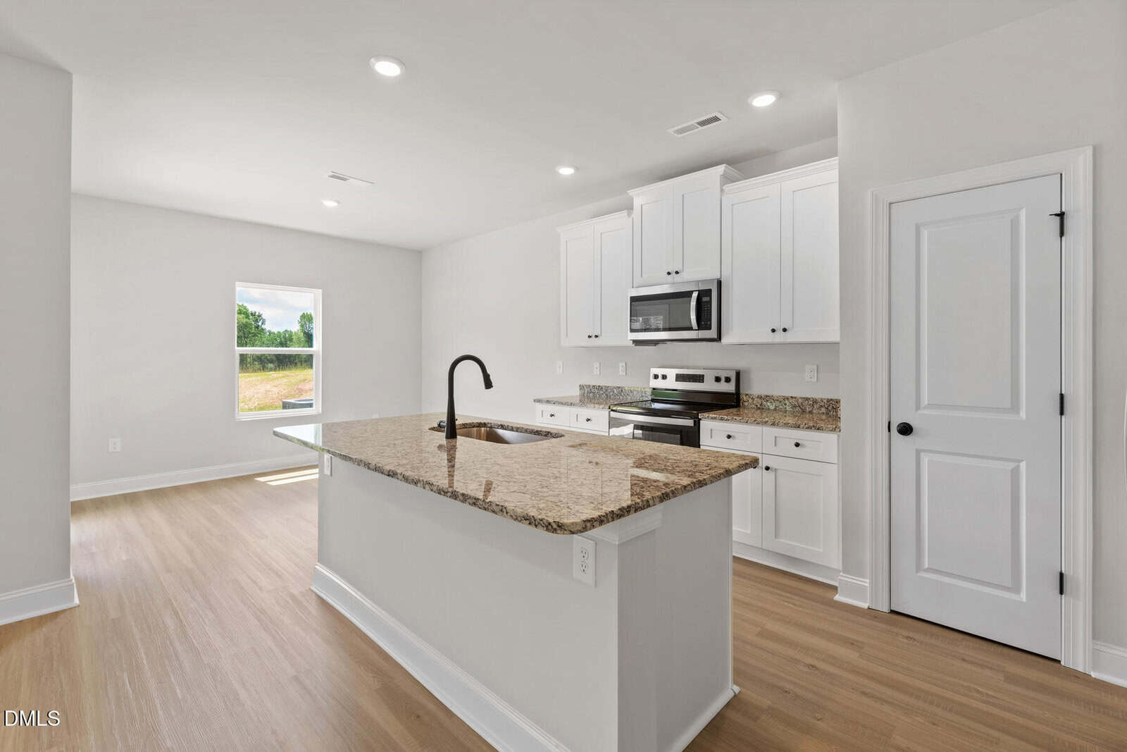 Modern kitchen with white cabinets, a center island featuring a granite countertop, built-in sink with a dark faucet, stainless steel appliances, light wood flooring, and a window letting in natural light.