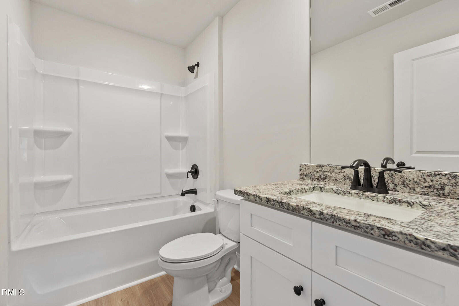 Modern bathroom with white bathtub and shower combo, white toilet, and white vanity with granite countertop and black fixtures. Light wood flooring and large wall mirror enhance the bright, clean look.