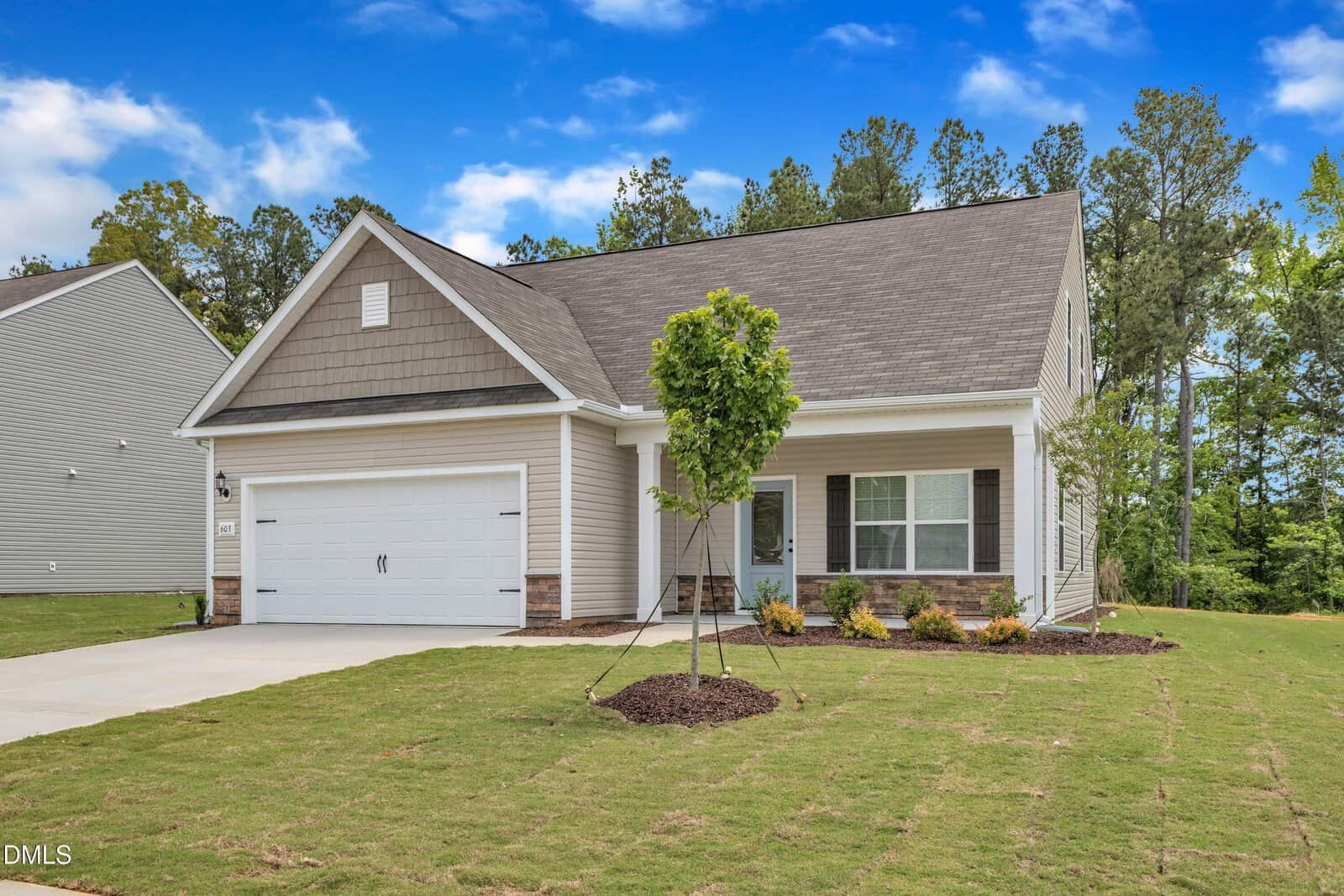 A single-story suburban house at 201 Benton Drive with a two-car garage, light-colored siding, and a gable roof. The front yard features new grass, a small tree, and minimal landscaping. Trees frame the home under a blue sky.