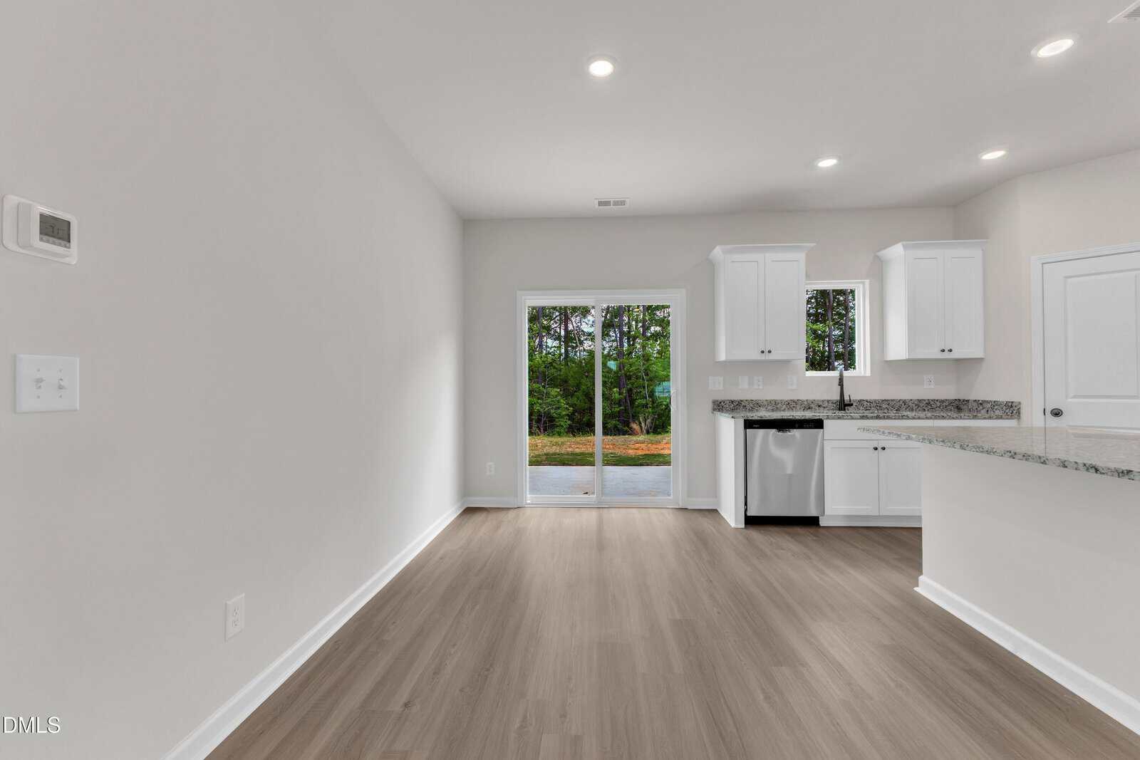 A modern, unfurnished kitchen and dining area with white walls, recessed lighting, wood-style flooring, white cabinets, granite countertops, and sliding glass doors leading to a backyard with trees.