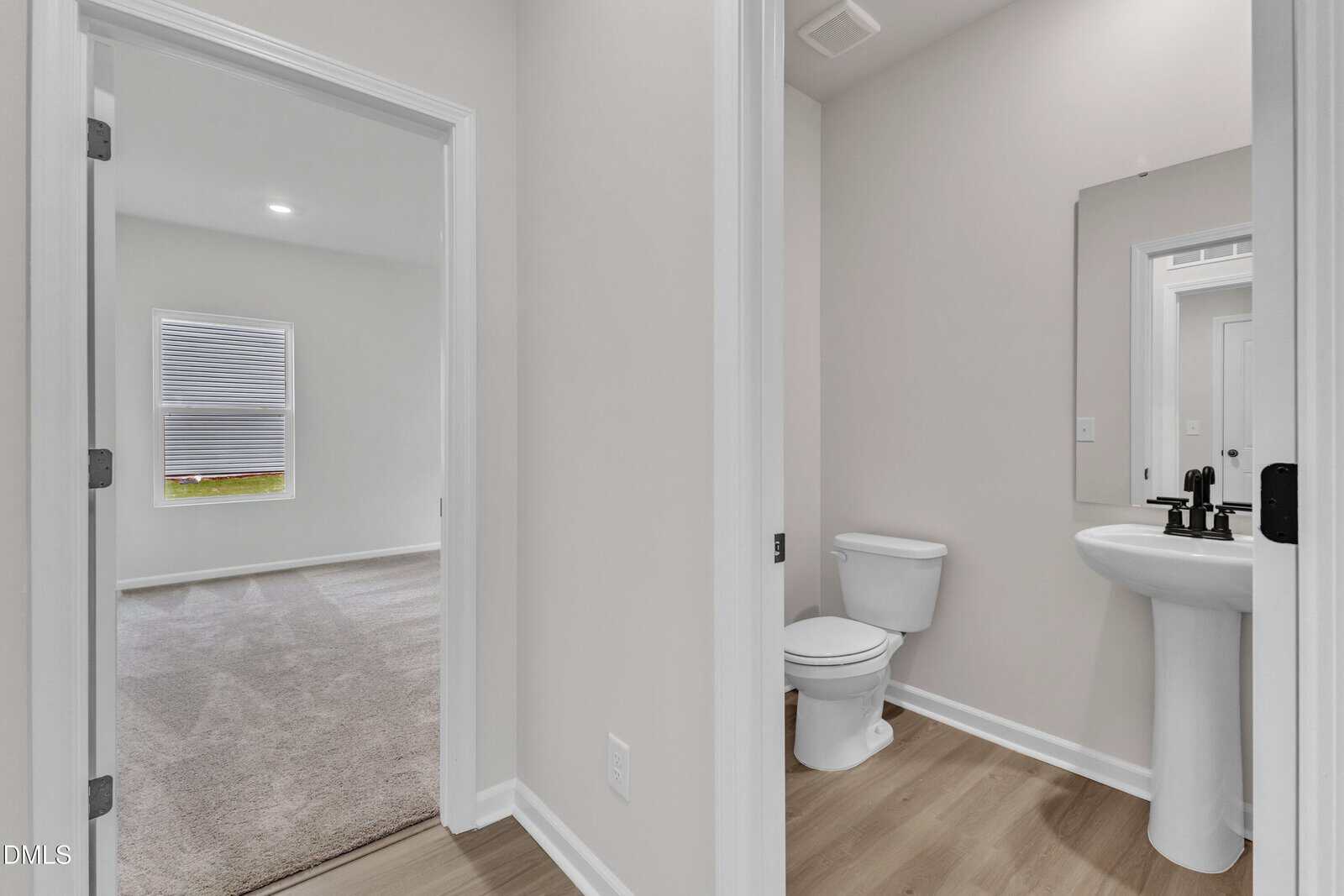 A view of a small bathroom with a white toilet, pedestal sink, and mirror, next to a hallway leading to a carpeted room with a window. The flooring in the bathroom and hallway is light wood.
