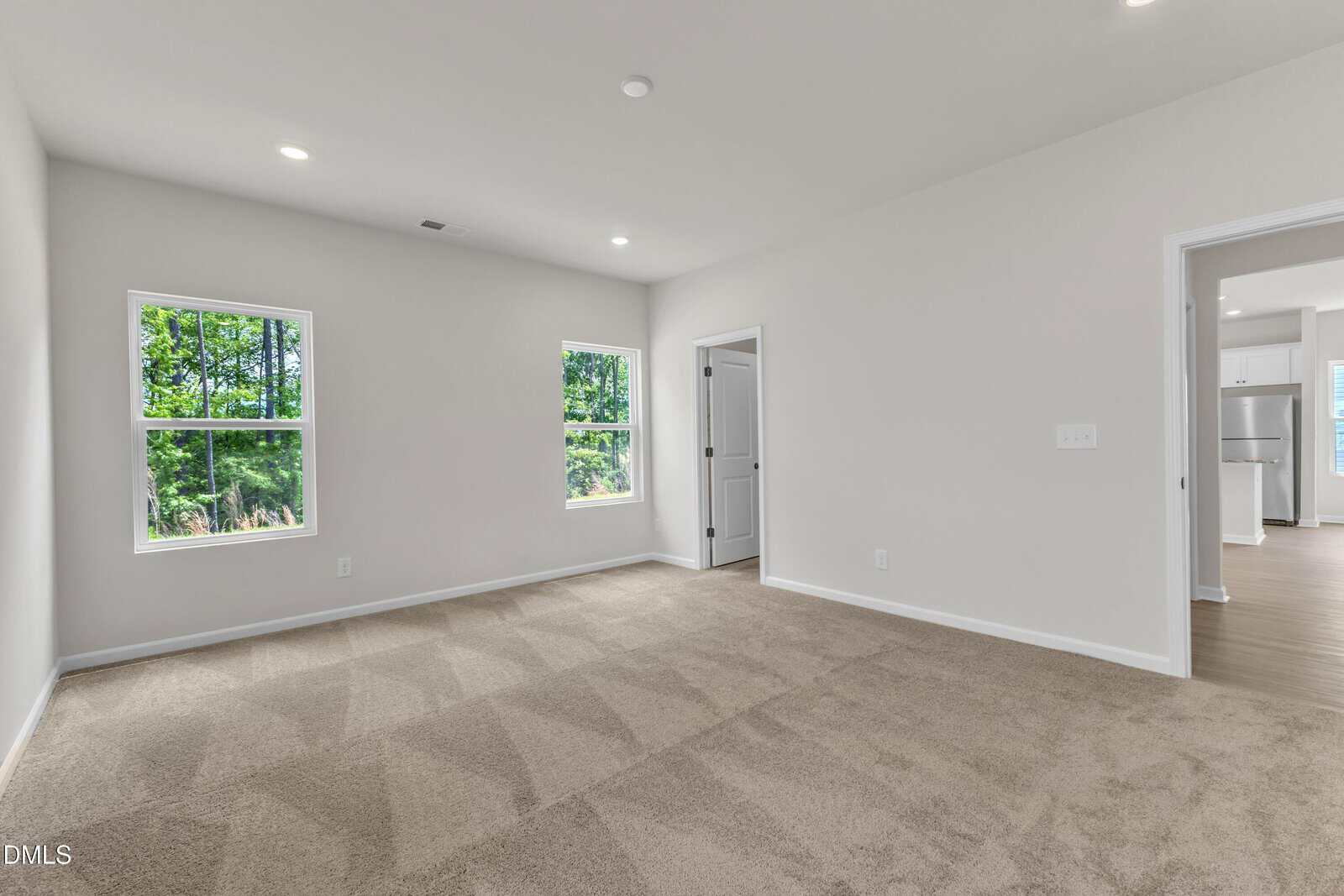 A spacious, empty room with beige carpet, two windows showing greenery outside, white walls, and doorways leading to a bathroom and another room with wood flooring and a kitchen visible.