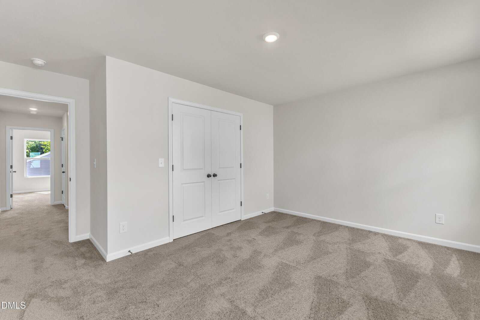 A bright, empty room with beige carpet, white walls, and double doors. An open doorway leads to another well-lit space with windows and carpeting. Ceiling lights provide additional illumination.
