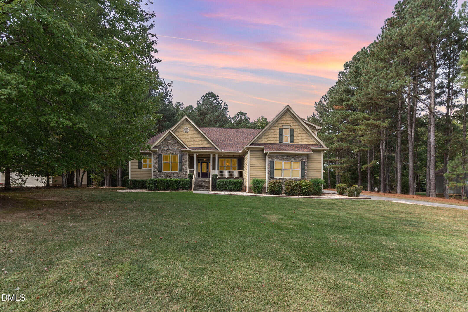 A yellow, single-story house with a front porch sits on a large grassy lawn, surrounded by tall trees under a colorful sunset sky.