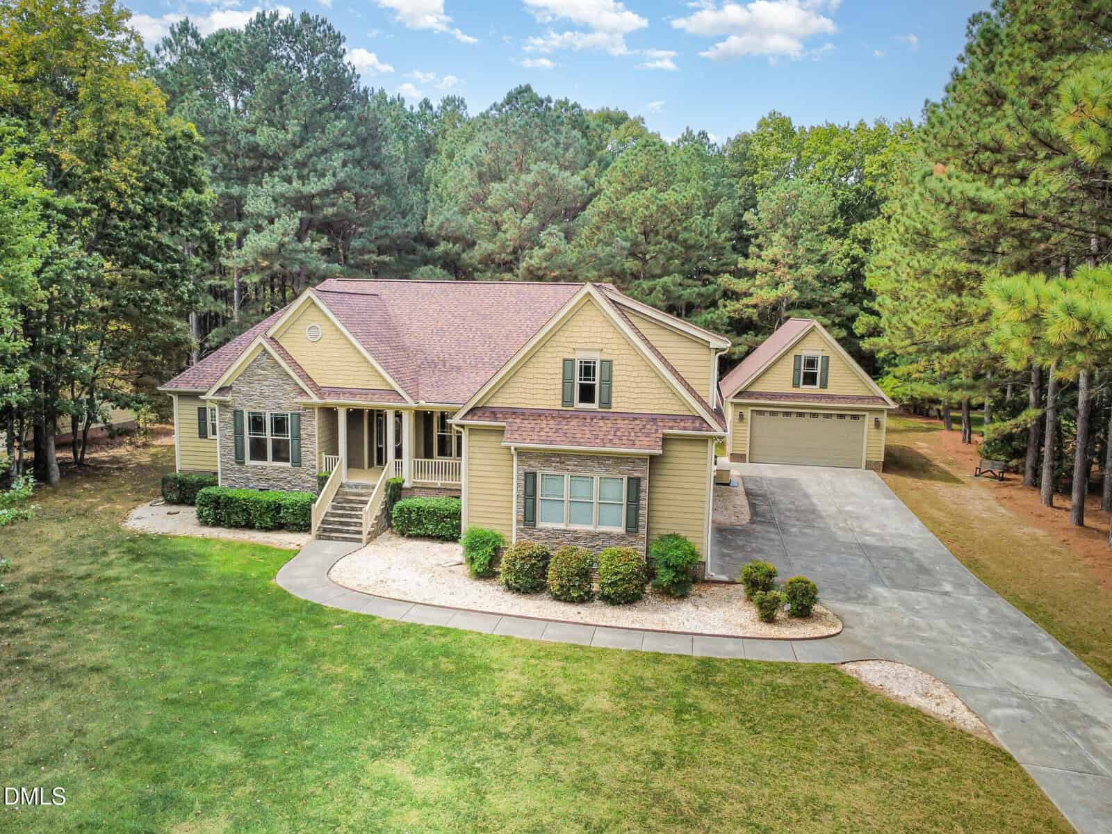 A large beige house with a front porch, surrounded by grass and trees, sits next to a detached two-car garage with a second story, connected by a wide driveway under a partly cloudy sky.
