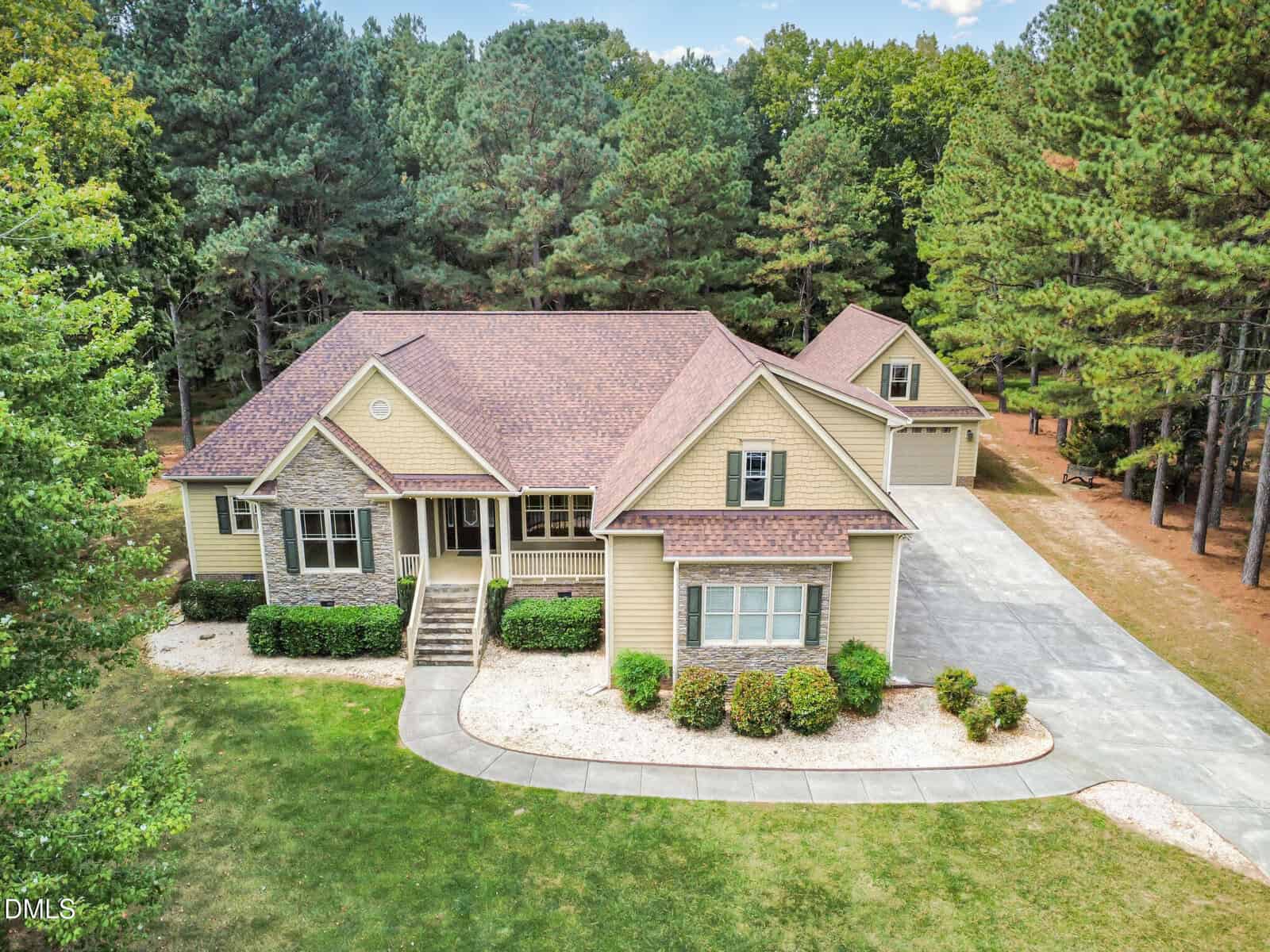 A beige house with a brown roof, front porch, and attached garage sits on a spacious lot with a curved driveway, surrounded by green lawn, shrubs, and tall trees. A detached garage is visible in the background.