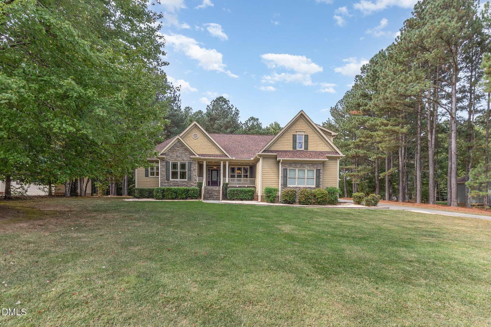 A tan suburban house with a covered front porch sits on a large, grassy lawn bordered by trees under a partly cloudy sky. A driveway runs beside the home, and landscaping lines the front walkway.