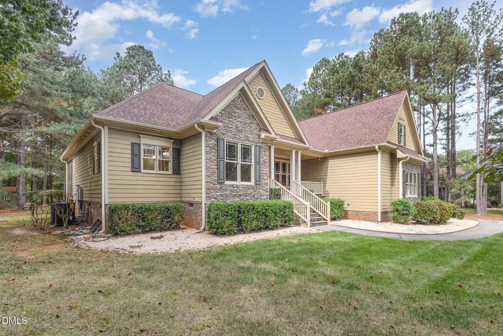 A beige house with a stone accent facade, gabled roof, and white trim sits in a grassy yard surrounded by trees under a partly cloudy sky. A small front porch with steps leads to the entrance.