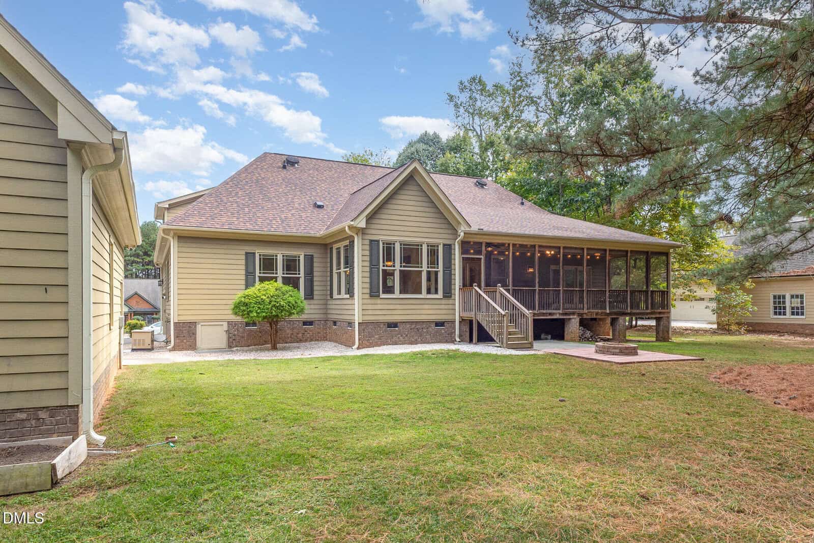 Back view of a beige house with a screened porch, wooden steps, small bushes, and a grassy yard, next to a detached garage under a partly cloudy sky.