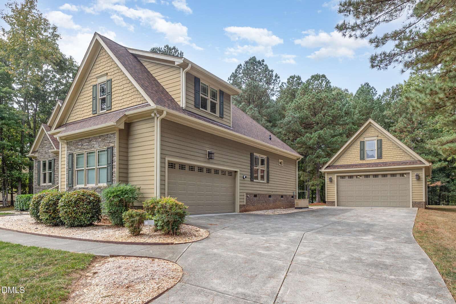 A two-story suburban house with beige siding, stone accents, and a three-car garage sits next to a separate matching garage. The property is surrounded by trees and has a curved driveway with landscaped shrubs.