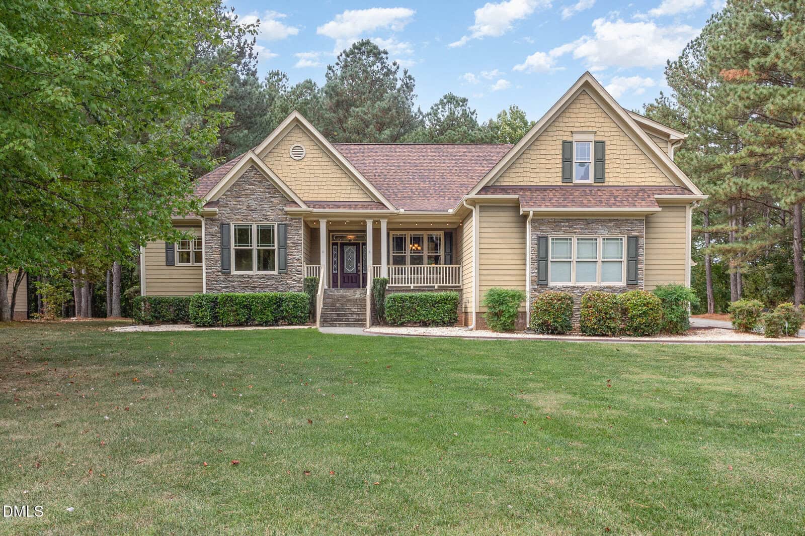A tan house with stone accents, front porch, and gabled roof sits on a large green lawn, surrounded by trees under a partly cloudy sky.