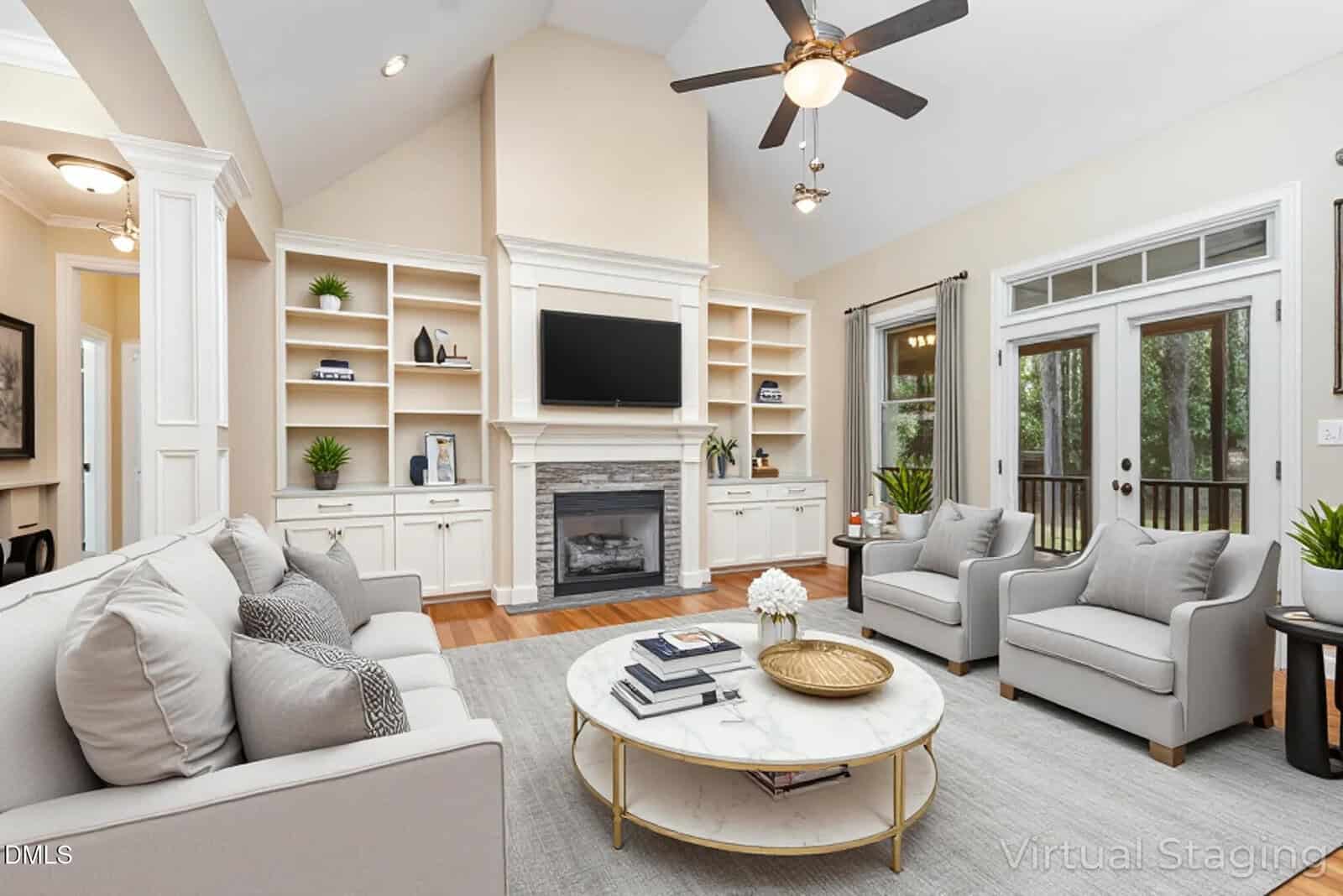 A bright, modern living room with light-colored sofas, two gray armchairs, a round marble coffee table, built-in shelves, a fireplace, and a TV above it. Large windows and glass doors let in natural light.