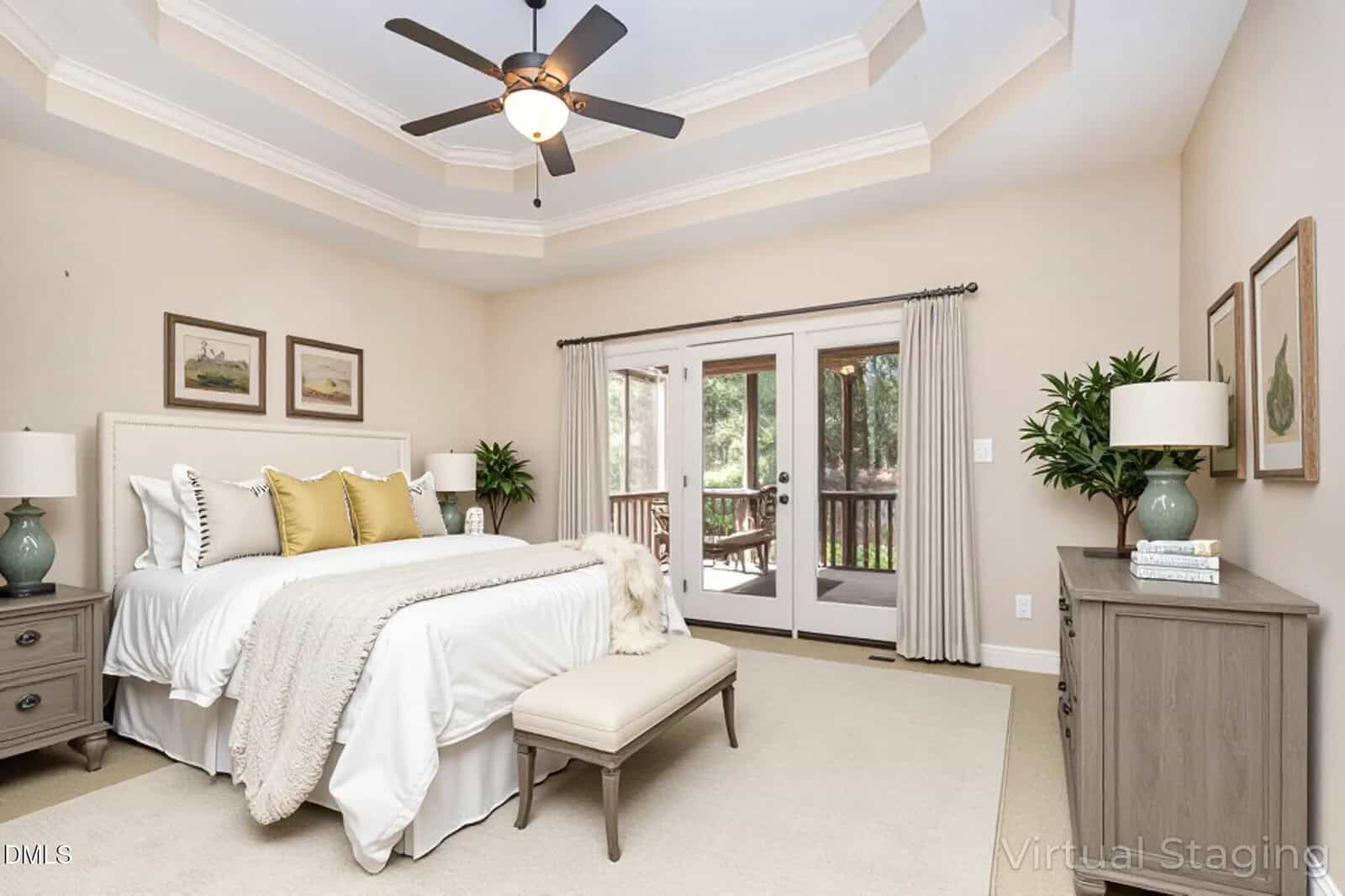 A spacious, neutral-toned bedroom features a neatly made bed with accent pillows, two nightstands, framed art, a ceiling fan, double doors with curtains, and a bench at the foot of the bed. Large windows show greenery outside.