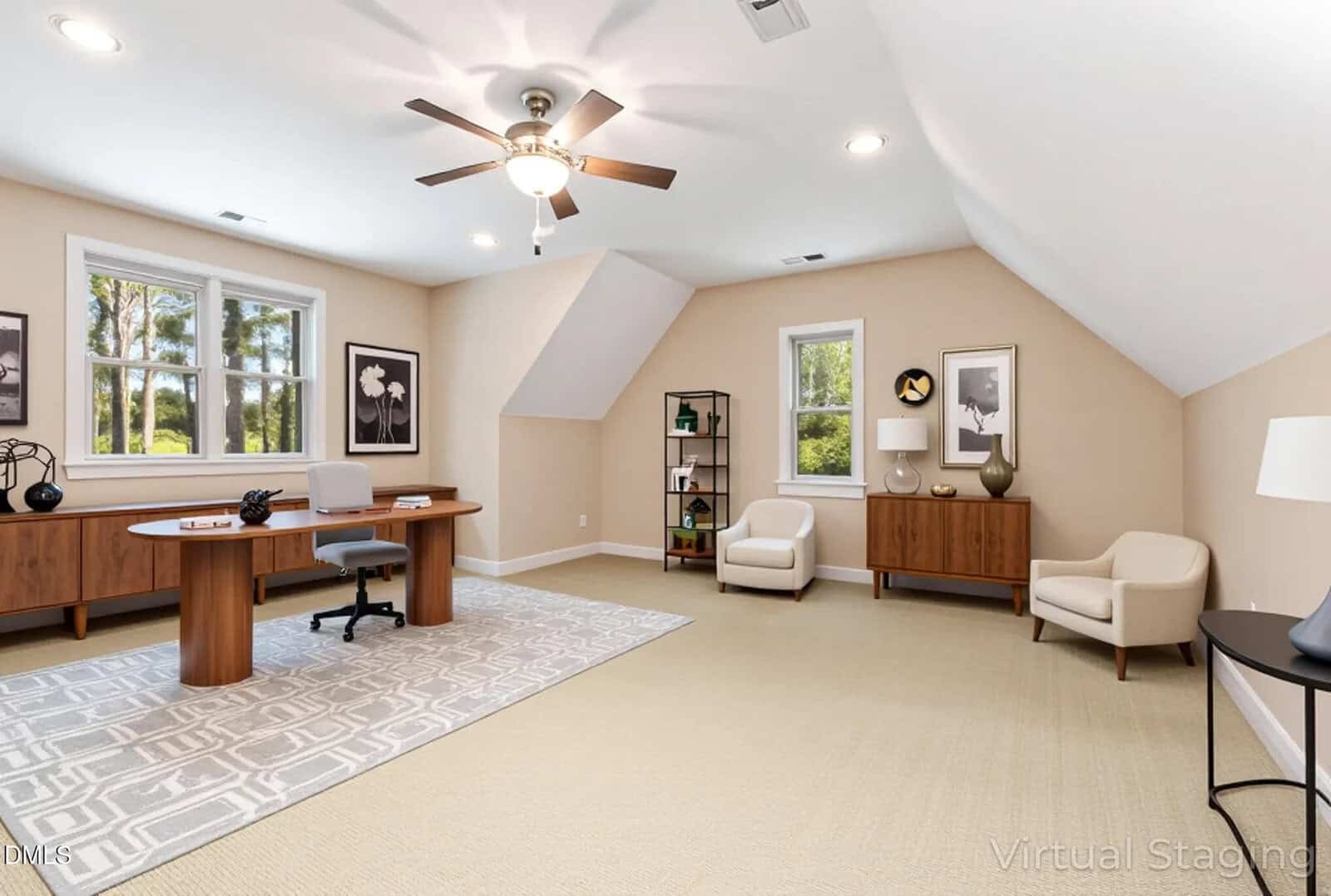 Spacious home office with a large wooden desk, office chair, armchairs, cabinets, wall art, and a ceiling fan. Room has beige walls, carpeted floor, and large windows letting in natural light.