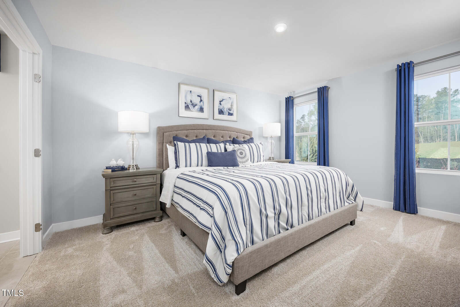 A bright bedroom with a large bed featuring blue and white striped bedding, two nightstands with lamps, framed art above the bed, beige carpet, and two windows with blue curtains letting in natural light.