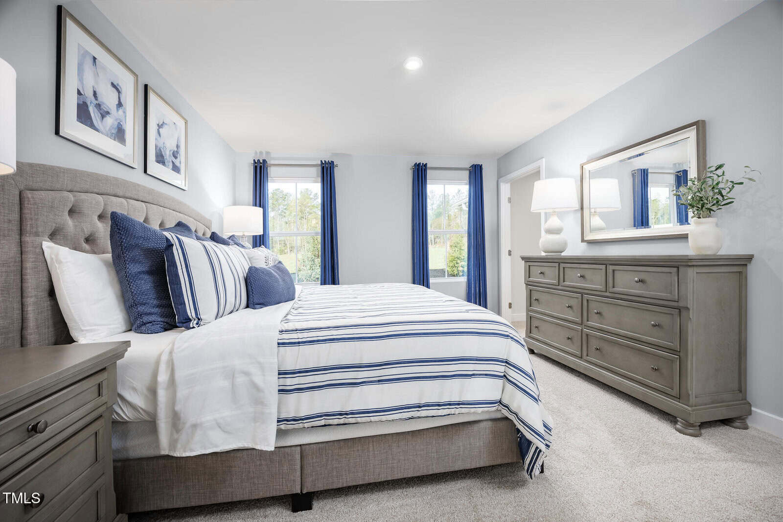 A bright bedroom with a neatly made bed featuring blue and white striped bedding, a tufted headboard, matching nightstands, a large dresser with lamps, a mirror, and two windows with blue curtains.