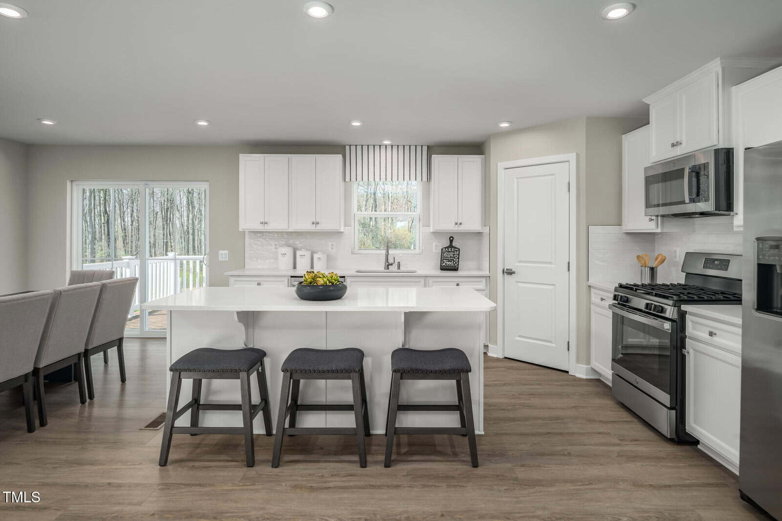 A modern kitchen with white cabinets, stainless steel appliances, a center island with three stools, wood flooring, and a dining table partially visible to the left. Large windows let in natural light.