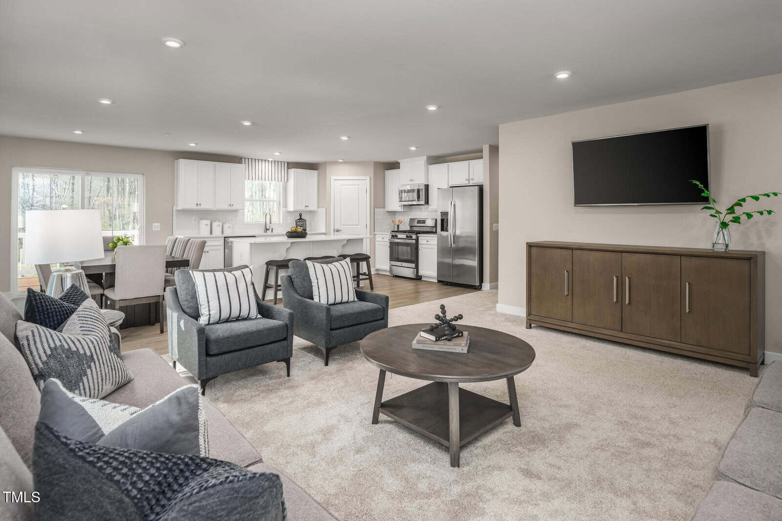 A modern open-concept living room with beige walls and carpet, gray sofas and armchairs, a round coffee table, a large flat-screen TV on a wooden cabinet, and a view of the kitchen with white cabinets and stainless steel appliances.