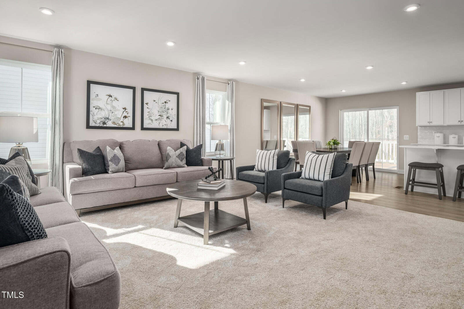 A spacious living room with neutral-toned furniture, including two sofas, two armchairs, and a round coffee table. The area opens to a dining space and kitchen, with large windows letting in natural light.