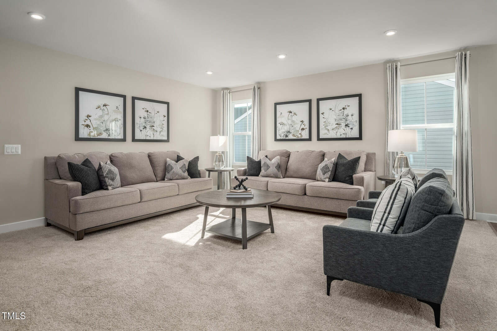 Modern living room with two beige sofas, two dark armchairs, a round coffee table, and three botanical art pieces on the wall. Light carpet and neutral decor create a bright, welcoming atmosphere.