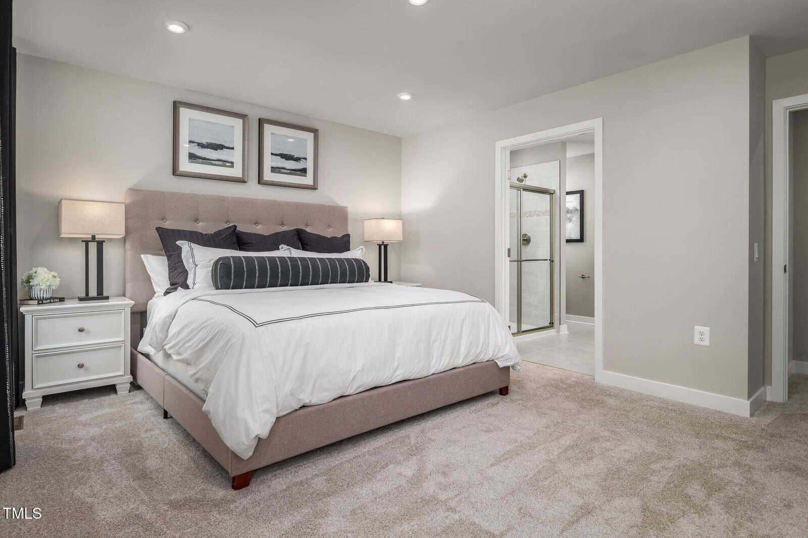 Modern bedroom with beige walls, a tufted bed with white bedding, two nightstands with lamps, and an open door leading to a bright bathroom with glass shower. Carpeted floor and framed art above the bed.