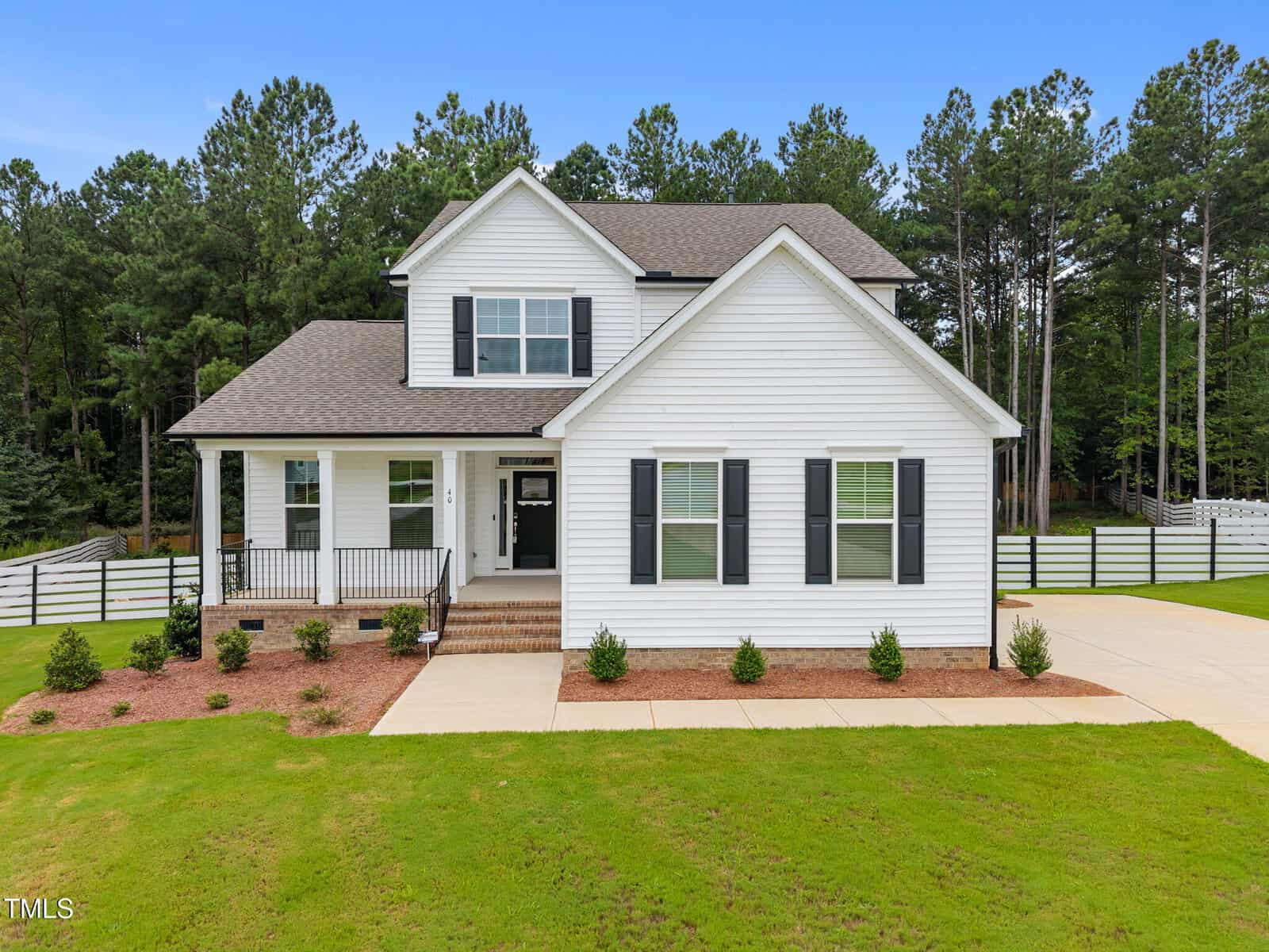 Charming two-story white house with black shutters, a covered front porch, and driveway located at 40 Long Needle Court in Youngsville. The lawn is green with small shrubs, and there are tall trees and a white fence in the background under a blue sky.