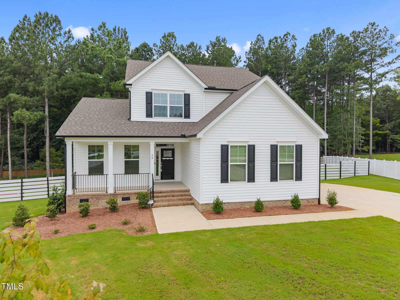 Two-story white house with black shutters, a covered front porch, and a driveway. The home is surrounded by a lawn with small shrubs and is bordered by a white fence and tall trees in the background.