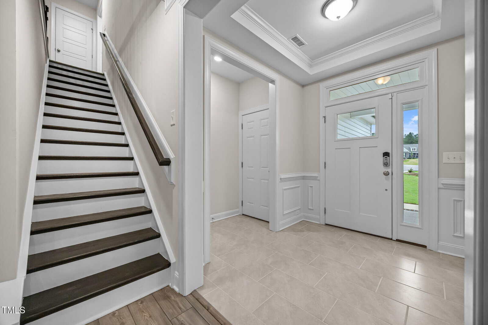 A bright entryway with a white front door, sidelight window, tiled floor, stairway with dark wooden steps, and hallway with double closet doors. Crown molding and white walls give a clean, modern look.