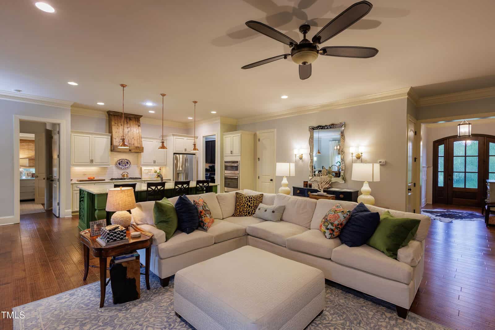 A spacious, open-concept living room features a large sectional sofa with colorful pillows, a cushioned ottoman, hardwood floors, and a view into a bright kitchen with pendant lights and an island.