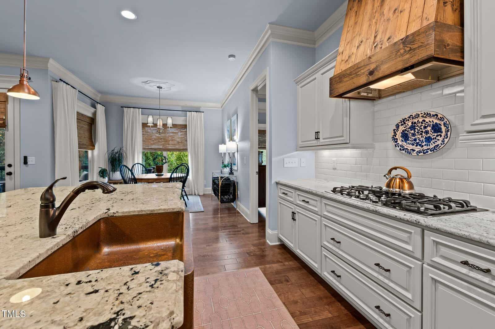 Bright kitchen with light granite countertops, a copper farmhouse sink, white cabinets, a wooden vent hood, a gas stove, and hardwood floors. The kitchen opens to a dining area with large windows and white curtains.