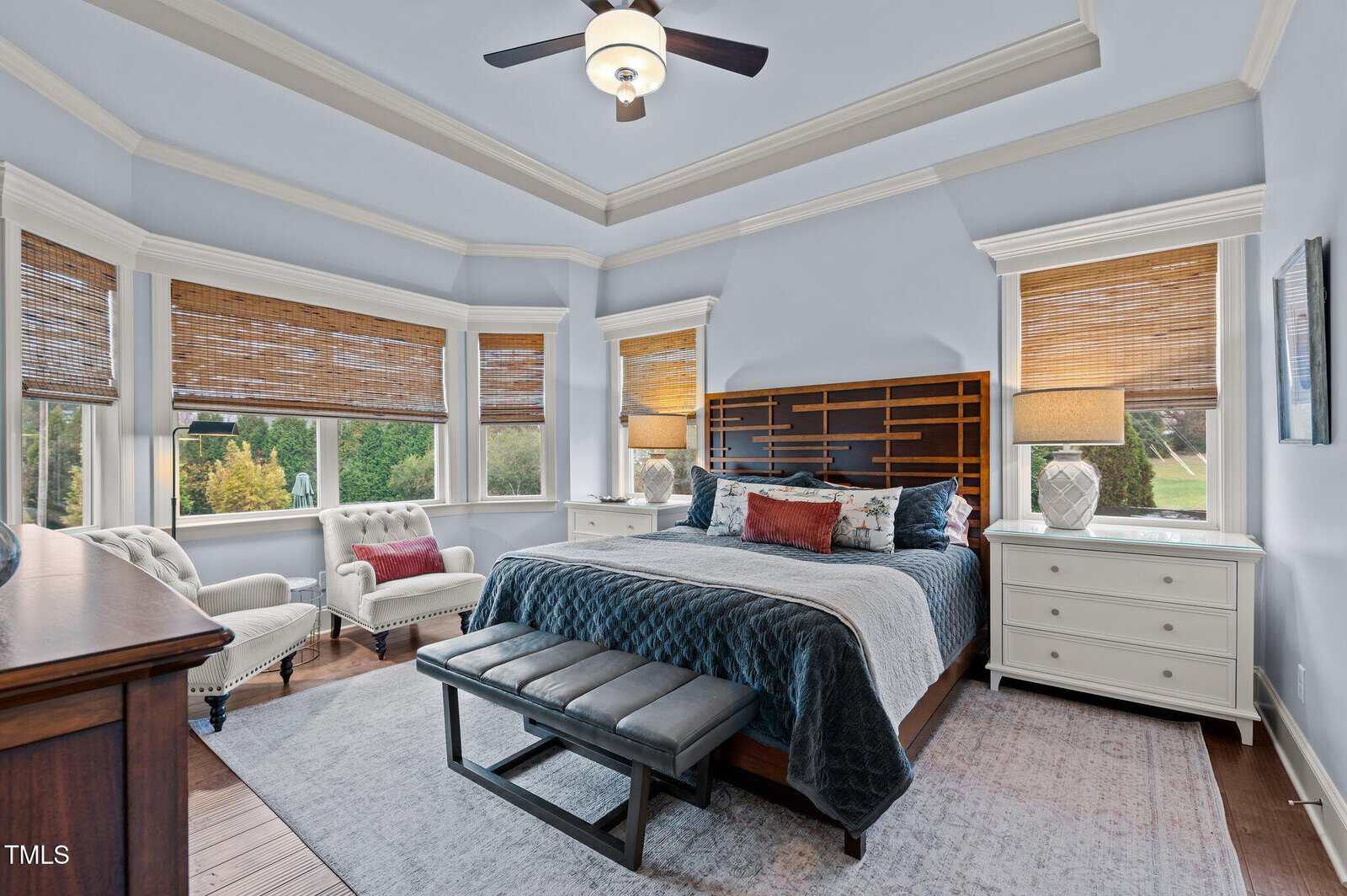 A spacious, light-filled bedroom with a tray ceiling, large windows with bamboo shades, a king-sized bed with a dark wooden headboard, two white armchairs, white nightstands, and a dark bench at the foot of the bed.