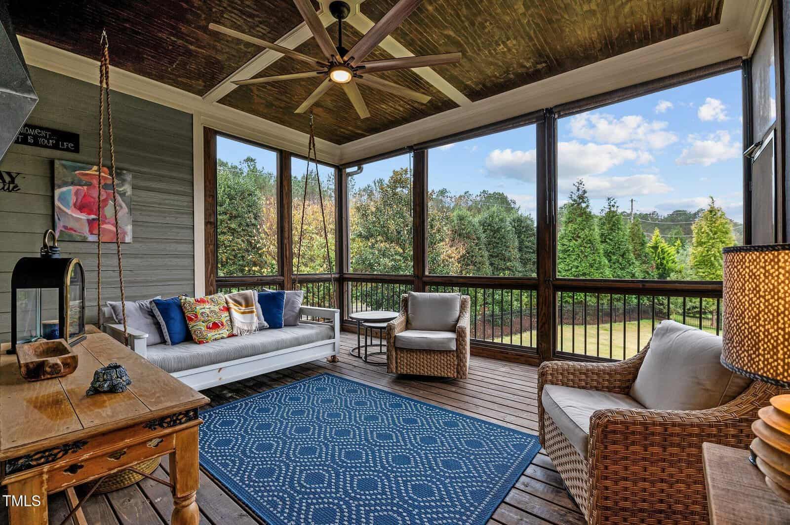 A cozy screened-in porch with a white sofa, two wicker armchairs, a blue patterned rug, a wooden table, and a ceiling fan. Large windows overlook a green yard with trees under a blue sky.