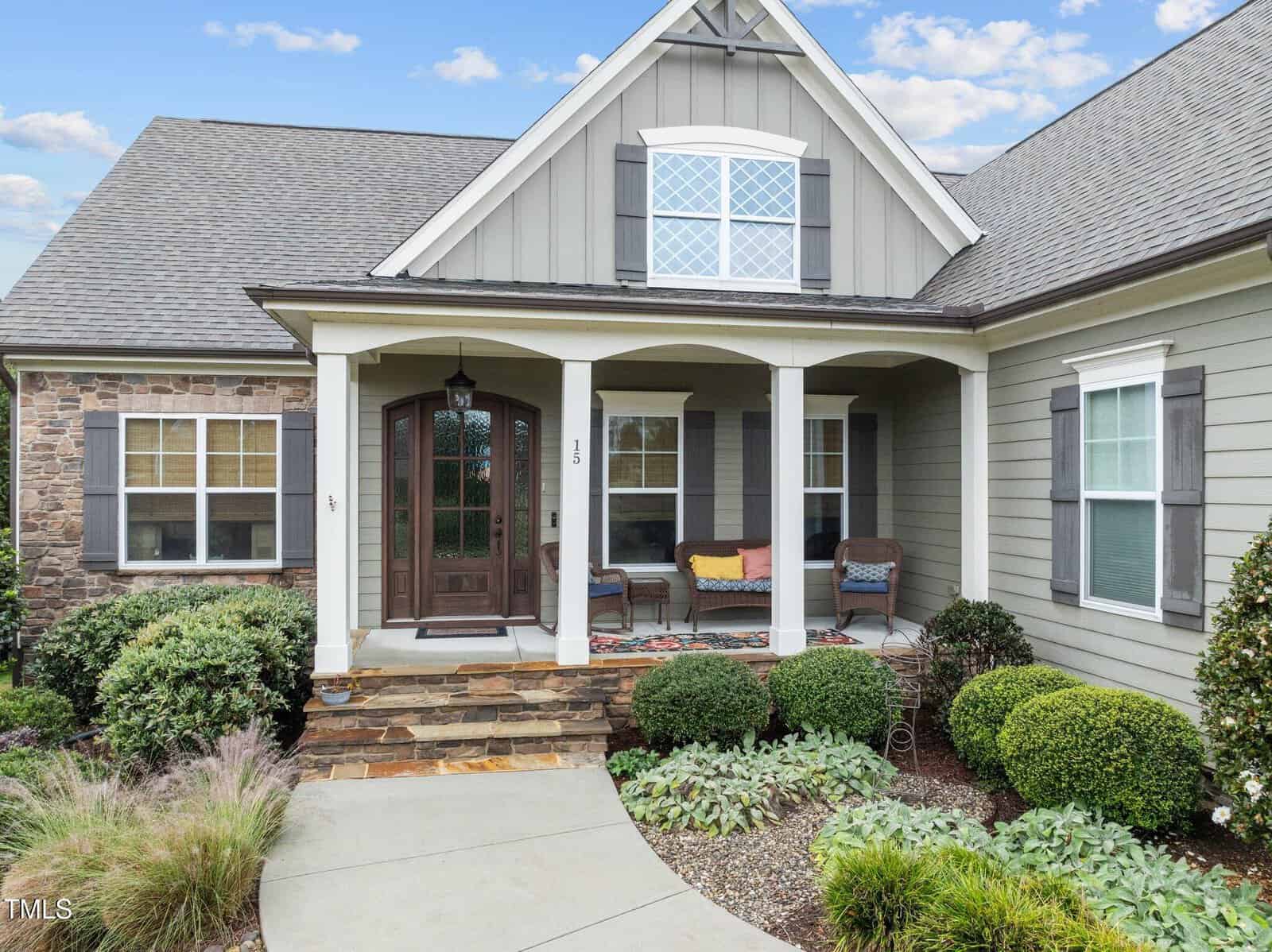 A charming house with gray siding, stone accents, and a covered front porch. The porch has two chairs, a bench with colorful cushions, and is surrounded by neatly trimmed bushes and landscaping.