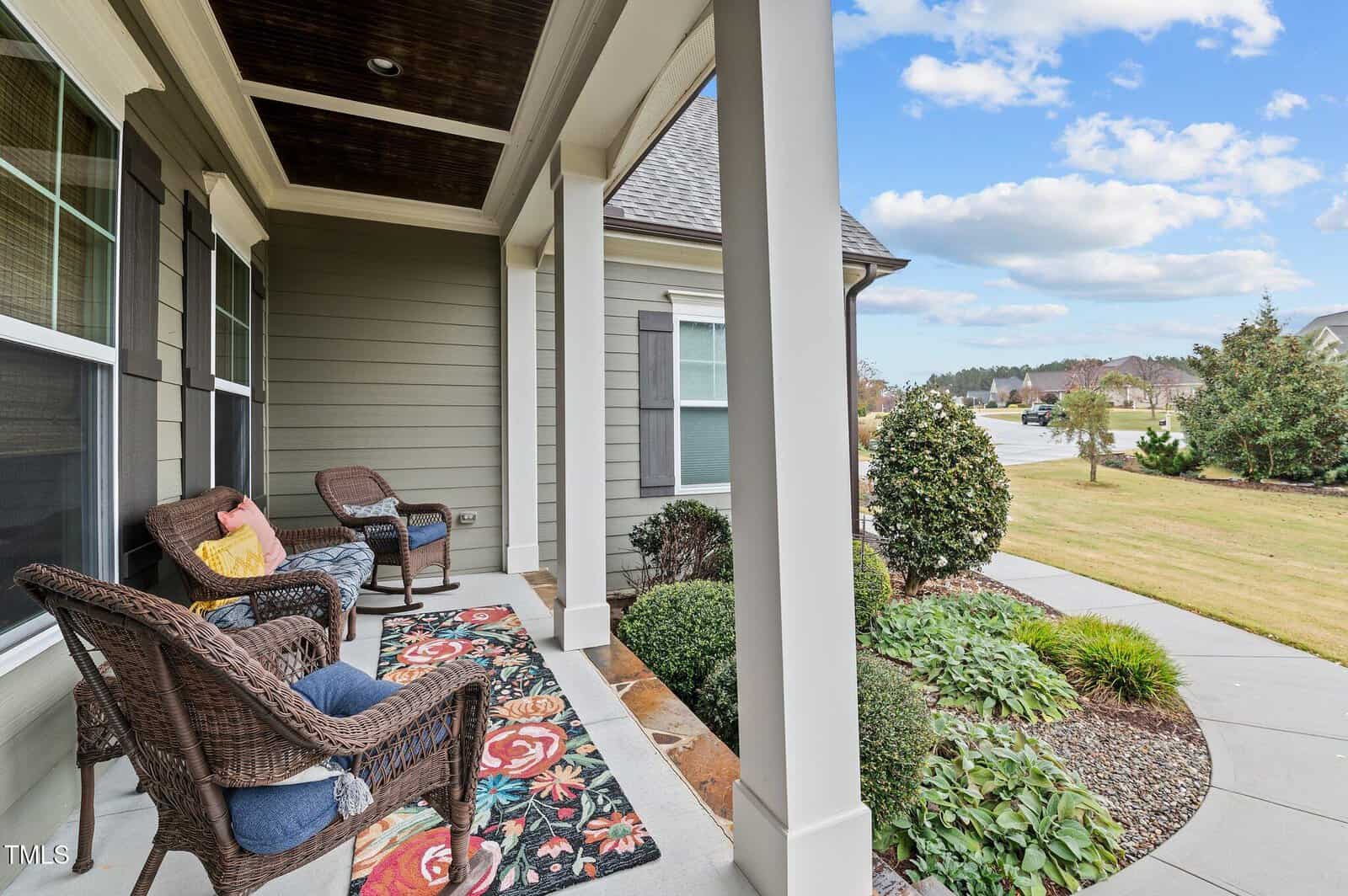 A cozy front porch with wicker chairs, colorful cushions, and a patterned rug overlooks a walkway, lawn, shrubs, and a suburban neighborhood under a bright, partly cloudy sky.