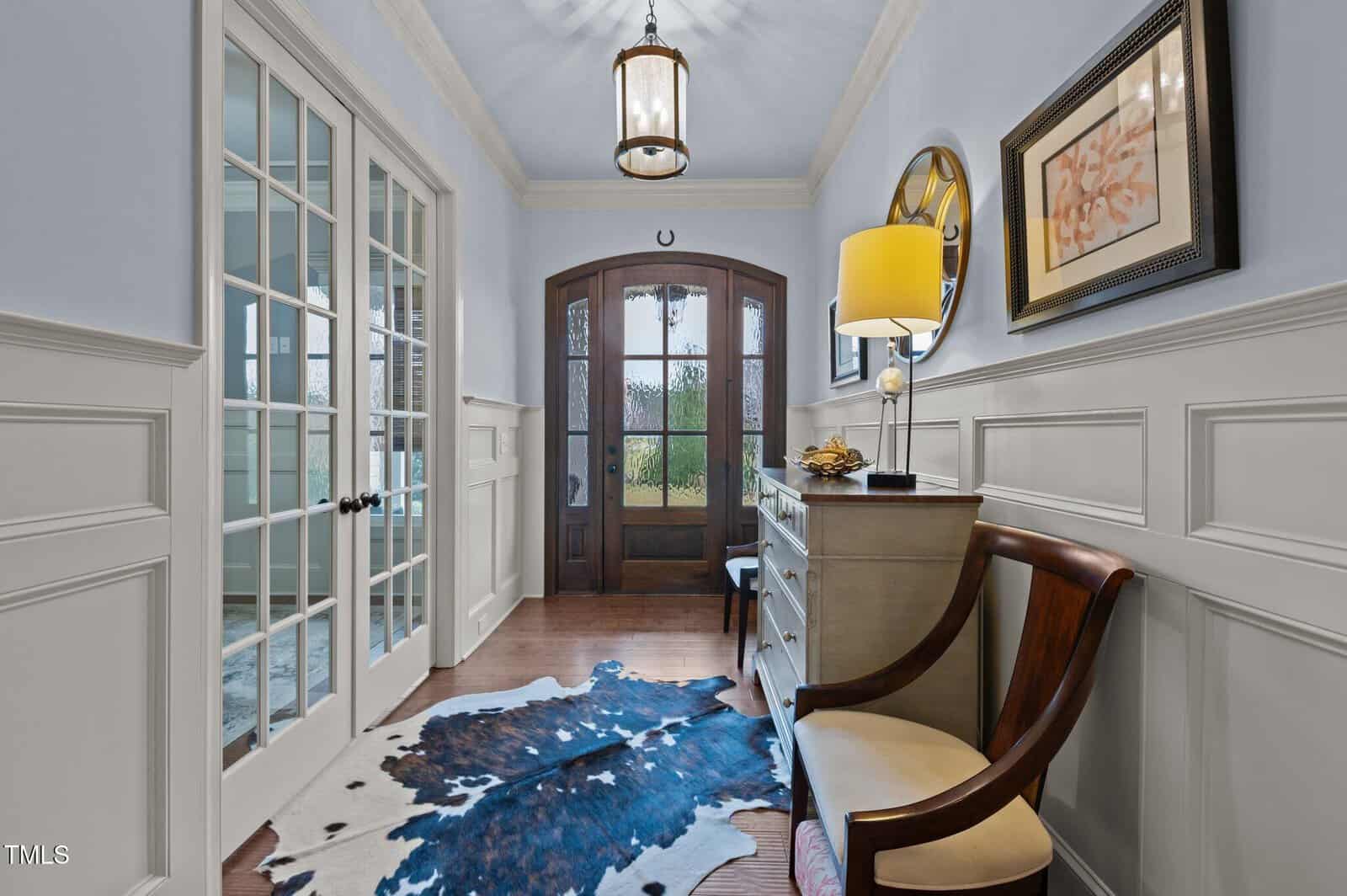 A hallway with wainscoting, a wooden chair, dresser with a lamp, cowhide rug, framed art, and double glass doors leading to another room. A pendant light hangs from the ceiling above the entrance.