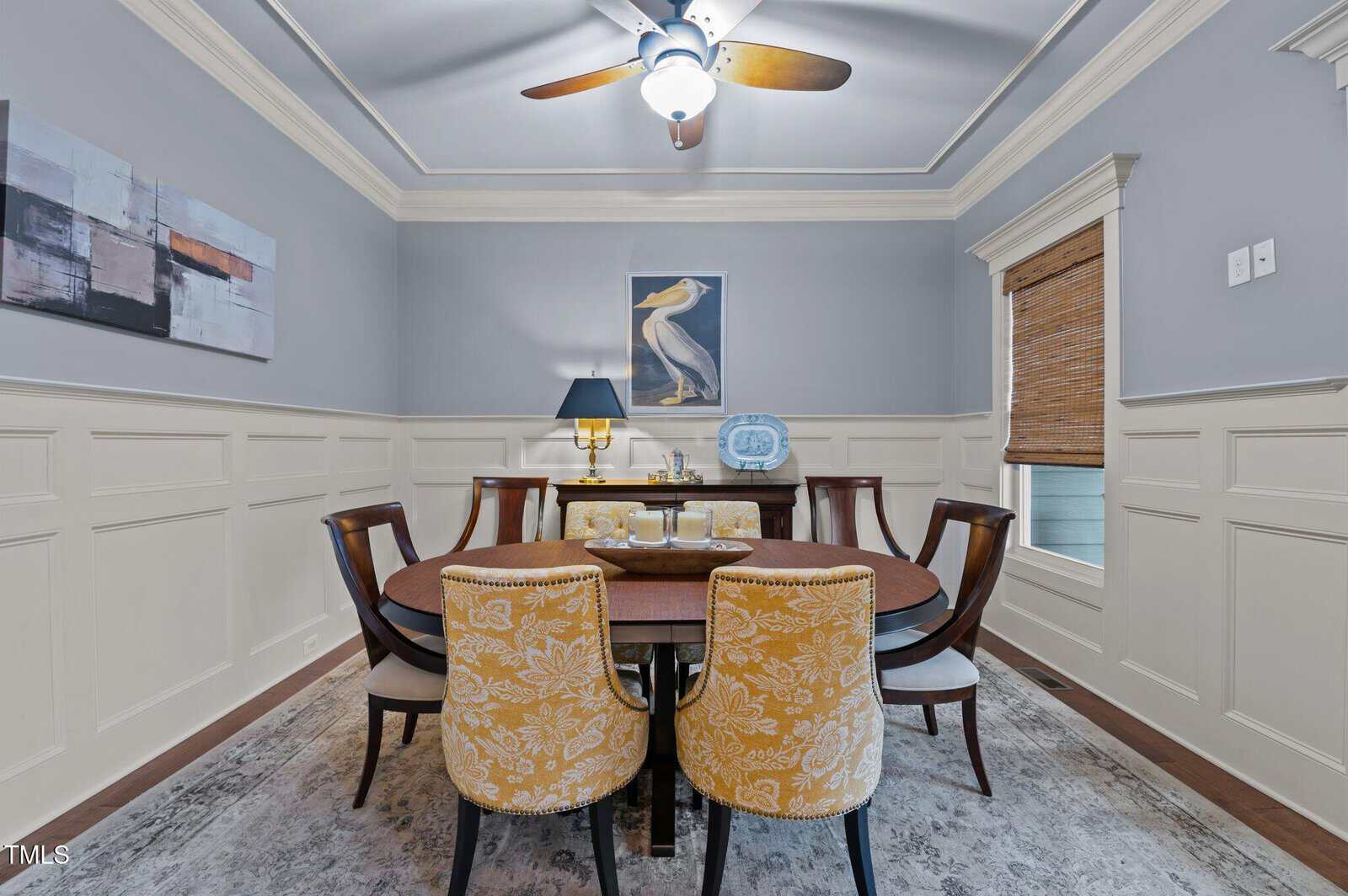 A dining room with a round wooden table, six chairs—two with yellow floral upholstery—art on the pale gray walls, a window with a bamboo shade, a ceiling fan, and a sideboard with a lamp and decorative plate.