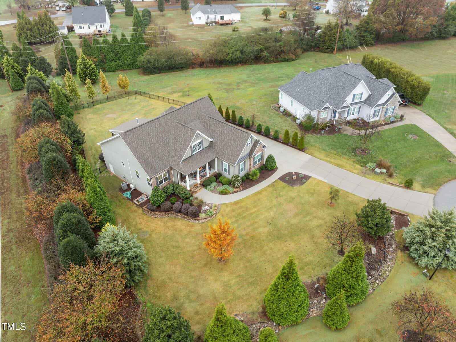 Aerial view of two suburban homes with gray roofs, surrounded by neatly landscaped lawns, shrubs, and trees. Curved driveways lead to each house, and the area is bordered by greenery and fields.