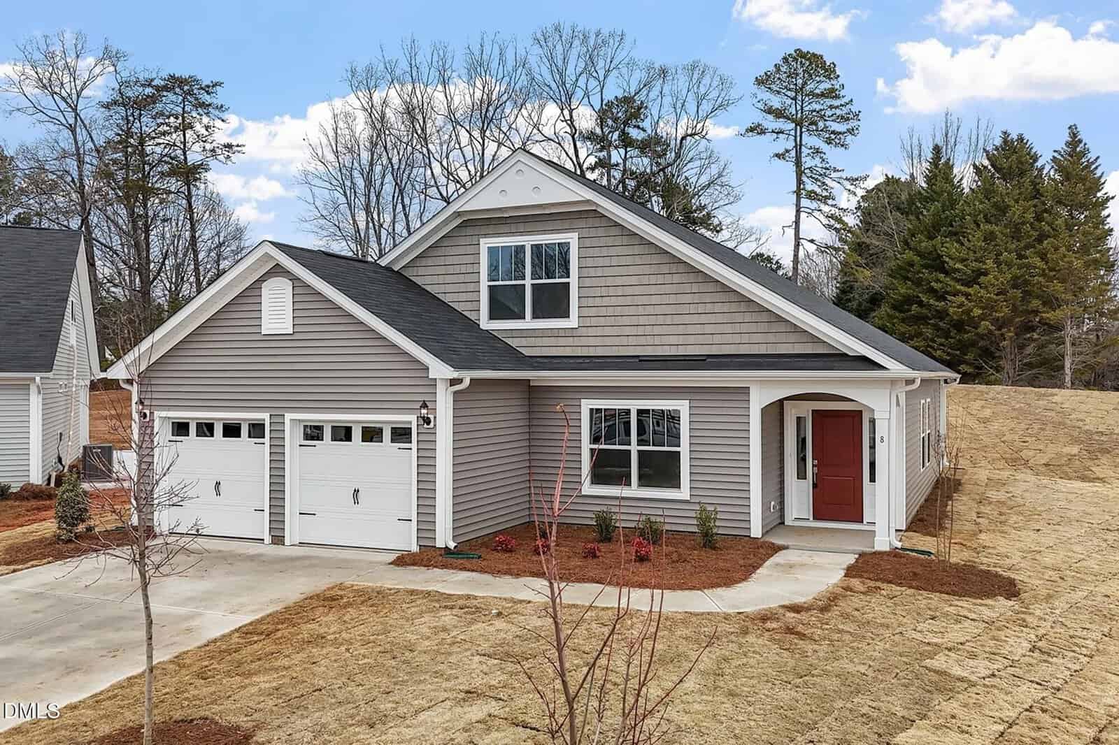 A single-story house with gray siding, two white garage doors, a red front door under a covered porch, and a small front yard with new landscaping and a concrete driveway. Leafless trees and pine trees are in the background.