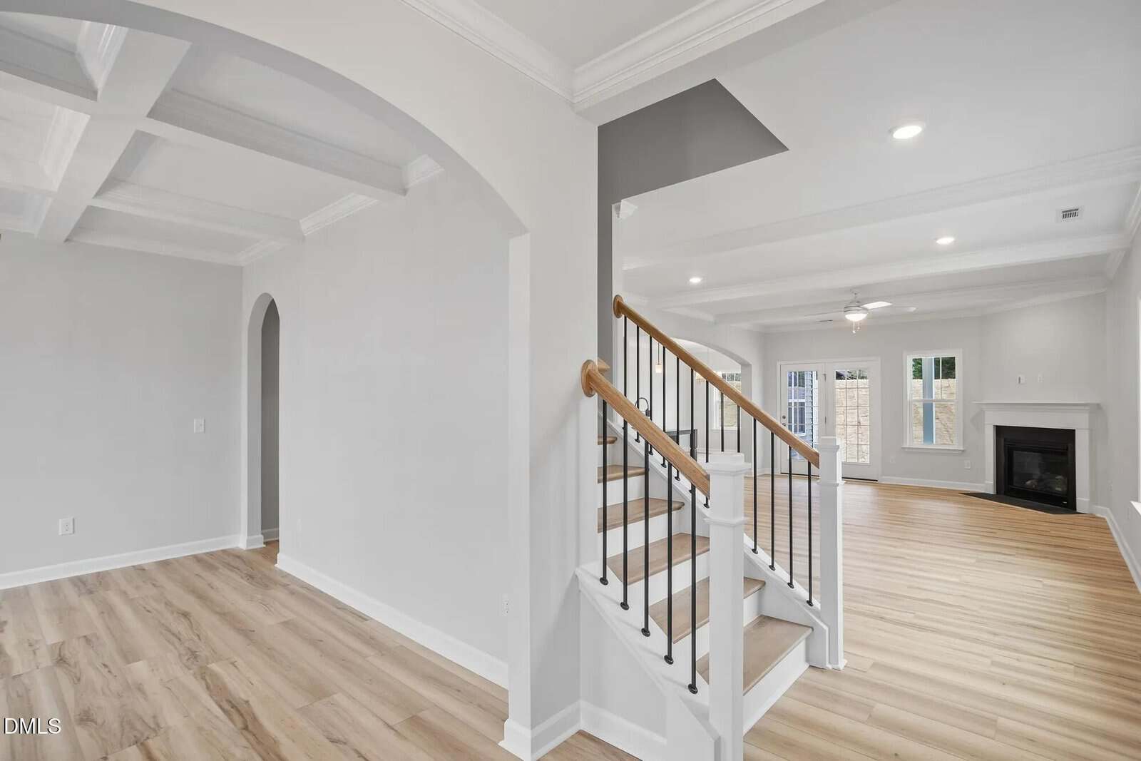 A bright, modern interior of a house featuring a staircase with wooden steps and white railings, light wood flooring, white walls, an archway, and a living area with a fireplace and large windows.