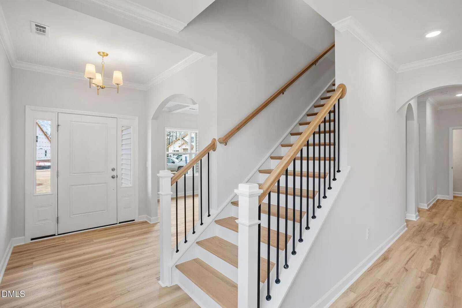 A modern entryway with light wood flooring, white walls, a white front door with side windows, a staircase with wood steps and black iron balusters, and a gold chandelier hanging from the ceiling.